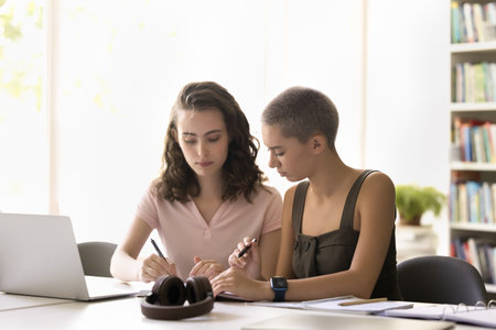 Helping friend. Focused hipster female student sit at desk study together with teen girl mate watch...