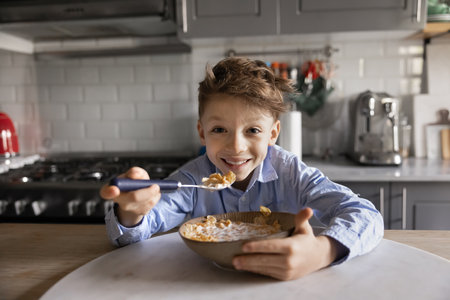 Cheerful kid having breakfast in home kitchen, looking at camera, smiling, posing for portrait. Funny cute early school aged boy eating corn flakes with milk, enjoying tasty sweet mealの写真素材