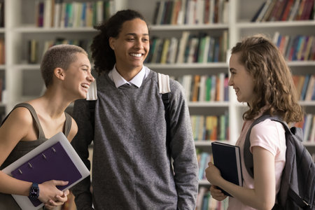 After lecture. Relaxed laughing multiethnic diverse students couple groupmates meet familiar teenage girl at public library stand together hold nice conversation talk to friend chat gossip having funの写真素材