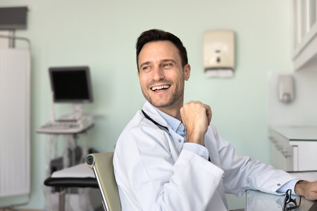 Cheerful handsome male doctor sitting at workplace, looking away with toothy smile, laughing, working at computer in ultrasound diagnosis room, having fun, enjoying job, medical careerの写真素材
