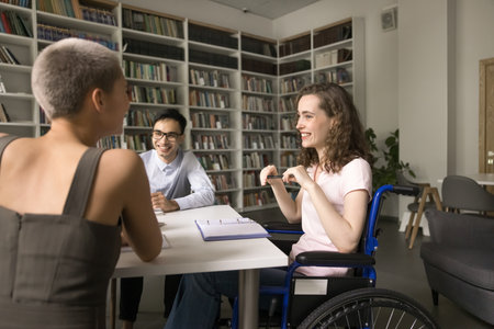 Learning equality. Smiling teen girl high schooler using wheelchair an inclusive class pupil student hold pleasant conversation share funny idea with laughing groupmates friends on workshop at libraryの写真素材