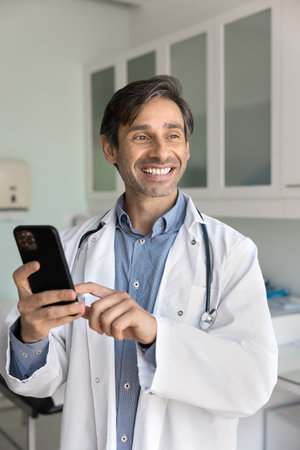 Happy middle aged Hispanic practitioner doctor man using mobile phone in his clinic office, typing, looking away, smiling, thinking on online communication with patients. Vertical shotの写真素材
