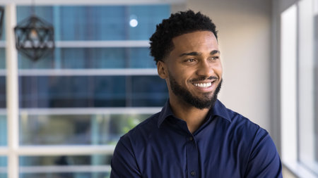 Happy dreamy African business leader man looking at window away with toothy smile, standing for casual portrait in office, thinking on successful career, leadership. Banner shot with copy spaceの写真素材