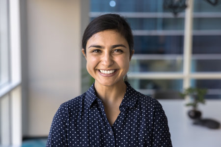Happy beautiful Indian businesswoman looking at camera with perfect toothy smile, posing for front head shot successful professional portrait, standing in office space aloneの写真素材