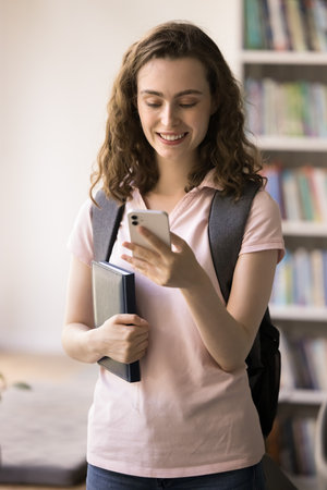 Vertical photography smiling confident teenage girl college university student stand at library using app on smartphone check book availability at online catalogue scroll news on high school web pageの写真素材