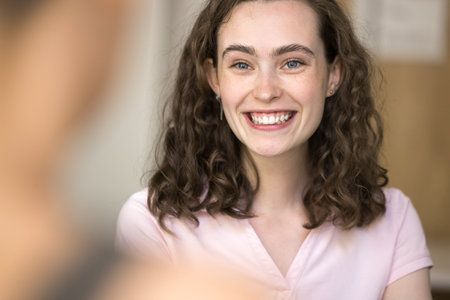 Pleasant conversation. Cropped portrait beautiful young lady with long curly hair talking with blurred person indoors hold nice dialogue enjoy discussion listen to partner with white smile. Copy spaceの写真素材