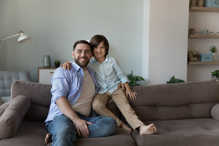 Loving preschooler son hugging young father sit on sofa smile feel happy enjoy leisure together at modern home, pose shooting for camera. Two generation people, good friendly relations, love conceptの写真素材