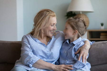 Middle-aged grandmother hugs her little granddaughter sit together on sofa, laughing enjoy conversation, joking having pleasant warm talk at home. Multi generational family ties, understanding, loveの写真素材