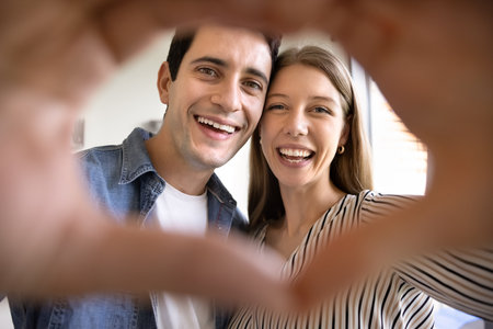 Cheerful Latin young dating couple in love looking at camera with toothy smiles through hand heart shaped frame, showing symbol of kindness, friendship, family, celebrating Valentines dayの写真素材