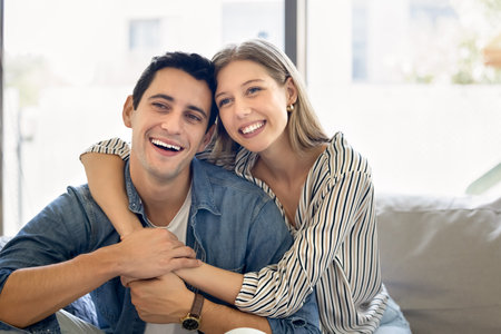Cheerful young Hispanic husband and wife hugging with love, sitting close on home sofa, looking away, smiling, laughing. Happy couple in love enjoying happiness in romantic relationshipの写真素材