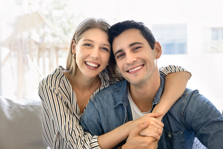 Happy young Latin couple in love hugging on home couch, looking at camera with perfect toothy...