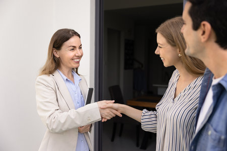 Positive confident realtor shaking hands with couple of young renters at entrance of rental apartment, smiling, finishing meeting. Happy customers giving handshake to agent, finding flat for rentの写真素材