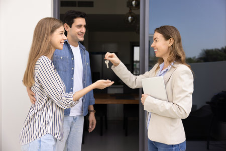 Confident female professional realtor woman giving key to couple of customers, offering rental to happy young husband and wife, meeting with new property owners at doorstepの写真素材