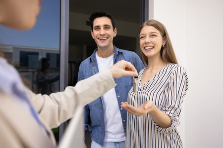 Happy couple buying new house, getting key from realtor, meeting with seller, real estate agent outdoors at apartment entrance. Positive young man and woman renting flatの写真素材