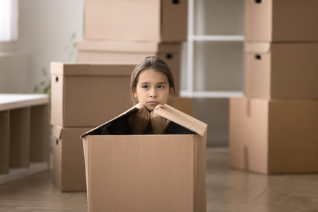 Serious pensive little girl sitting inside big cardboard box, surrounded by stacked boxes on relocation, looks bored monotony of move-out day, feeling of uncertainty or discomfort in new environmentの写真素材