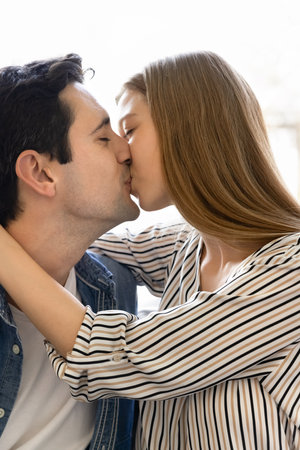 Happy young Hispanic couple in love kissing on lips indoors, hugging with closed eyes. Latin husband and wife enjoying romantic moment, close relationships. Vertical casual portraitの写真素材