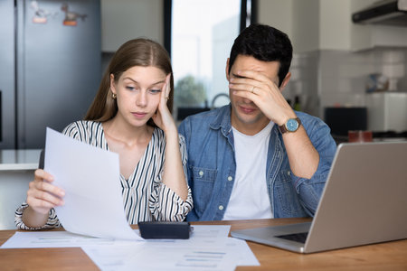 Concerned young couple of tenants, renters checking too high paper bills at home, sitting at laptop, calculator, counting expenses, thinking on money loss, bankruptcy riskの写真素材