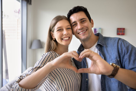 Happy attractive young couple making hand heart gesture together, joining fingers to form symbol of love, care, kindness, romance, looking at camera, smiling for portraitの写真素材