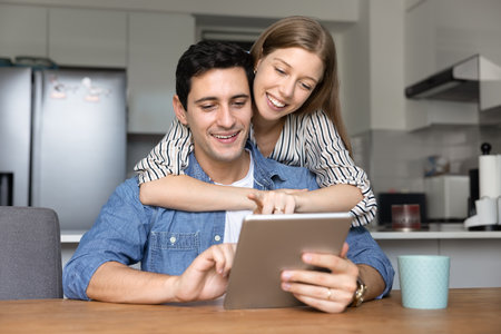 Happy young couple using digital tablet at home kitchen table, activating smart home virtual assistant, application, service, typing on gadget, enjoying modern wireless Internet domestic technologyの写真素材