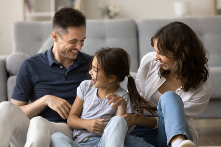 Young laughing parents tickling little cheery daughter seated together on floor in living room, feel overjoyed, enjoy funny playtime, priceless time and lively games together on family weekend leisureの写真素材