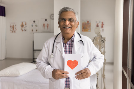 Cardio diseases prevention. Happy senior doctor physician cardiologist stand at modern cardiology clinic cabinet look at camera show you picture with read heart promote wellness regular health checkupの写真素材