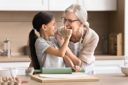 Candid laughing preteen child girl play at domestic kitchen with joyful granny having fun enjoy easy cooking food. Cute grandma and grandkid joking when preparing homemade pie paint noses in flourの写真素材