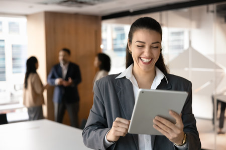 Excited cheerful young businessman using tablet in office meeting space, smiling, laughing, enjoying business success, career achievement, growth of sales, working on successful Internet projectの写真素材