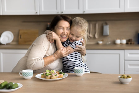 Granny and grandchild girl embracing in kitchen, showing affection, deep bond. Multigenerational family enjoy communication, cuddles, food they prepared together, eating sandwiches, talking, huggingの写真素材