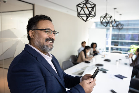 Cheerful mature Indian CEO man with smartphone standing in meeting room with team of employees, colleagues at large table, holding mobile phone, chatting, looking away, smiling, laughingの写真素材