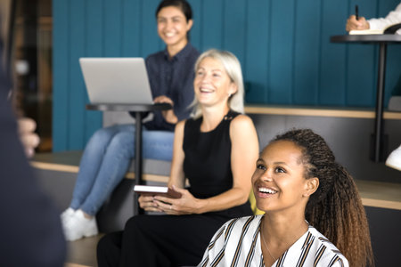 Group of diverse professionals seated in tiered, informal meeting space engaging in discussion with standing presenter. Presentation, team briefing, open communication, idea sharing, and collaborationの写真素材