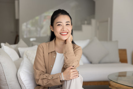 Happy positive young Asian girl posing on cozy sofa for home portrait, sitting on couch, leaning chin on hand, looking at camera, smiling, laughing. Vietnamese female homeowner girl head shotの写真素材