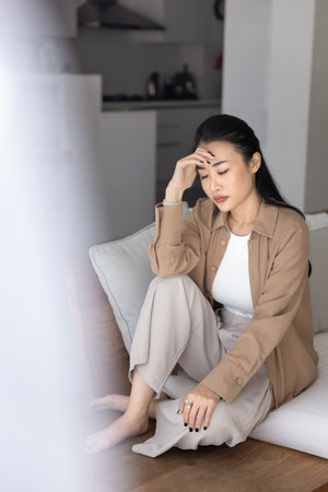Sad unhappy young Asian woman sitting on home couch, going through crisis, facing serious problems, grief, loss, depression, touching head, feeling headache. Vertical shot with cropped blurred veilの写真素材