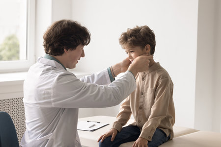 On pediatric examination. Attentive young male doctor family therapist gp see sick little boy at hospital cabinet touch young patient neck check inflamed lymph nodes to make diagnosis of flu influenzaの写真素材