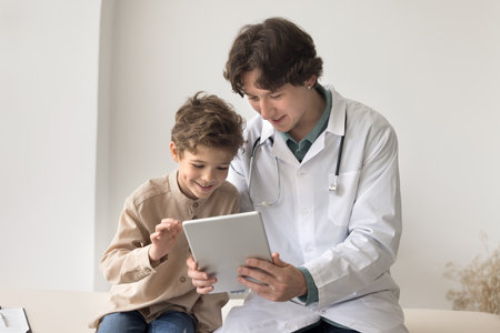 Caring young male doctor surgeon pediatrician use tablet pc on meeting with little boy play video...