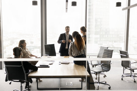 Five multiethnic staff members take part in morning briefing in high-rise office building behind closed doors, gathered together for teamwork, opinion sharing, strategizing and planning cooperationの写真素材