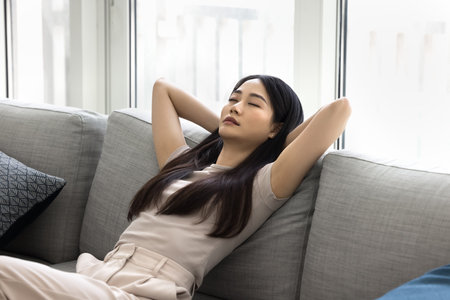 Calm Asian woman settled down on comfortable sofa with hands behind head, close her eyes daydreaming, relieving tiredness on carefree day-off alone at own or rented apartment. Stress free relaxationの写真素材