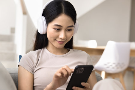 Close up of young Asian woman wear headphones using smartphone sitting comfortably on couch. Female listen to music or engaging in chat, immersed in audio messages sharing, enjoying pastime at homeの写真素材