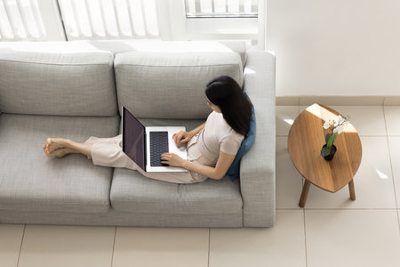 Woman sitting on cozy large sofa with laptop, focused on telework in relaxed posture, studying, browsing wireless internet, overhead top view. Remote work, leisure time using technology, productivityの写真素材