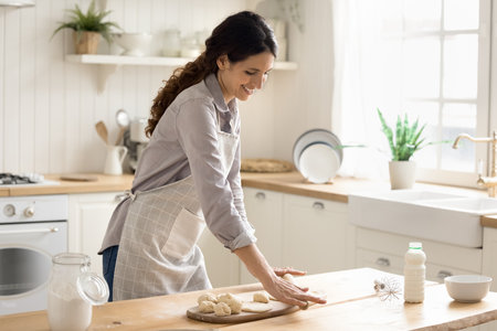 Home baking is my hobby. Smiling young woman housewife in apron rolling dough on kitchen table preparing homemade pies buns pastries for family breakfast enjoy cooking baked sweet dessert. Copy spaceの写真素材