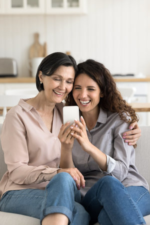 Family leisure time and digital tech. Laughing candid young daughter elderly mom cuddle on sofa at kitchen relax shoot funny selfie photo on mobile phone enjoy hilarious vlogs read funny message textの写真素材