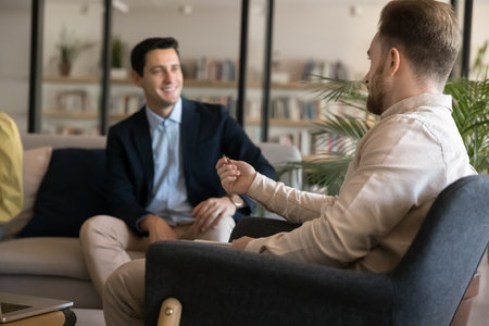 Casual conversation. Two young businessmen entrepreneurs students rest at library relaxation area hold informal dialogue chat sitting in comfy armchair sofa share ideas thoughts in friendly atmosphereの写真素材