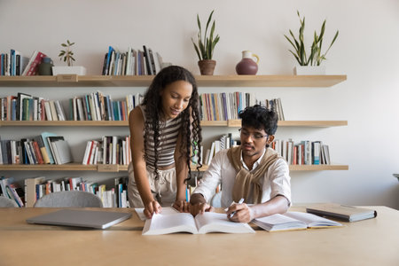 Two schoolmates or students doing university assignment, studying in college library, reading book together, researching, prepare essay or for exams, share knowledge. Mentorship, education, teamworkの写真素材