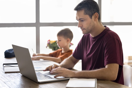 Young 35s focused man working on modern laptop while his little 7s school-age son drawing pictures, study or makes exercise seated together at table at home. Remote work, education, family lifestyleの写真素材