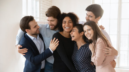 Six diverse workmates hugging indoors, gesture of camaraderie and celebration of achieving goal....
