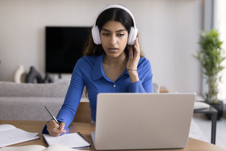 Focused Indian student woman sit at desk, wear wireless headphones, listen audio course, translate, jotting information, e-learns, improve language skills using laptop and educational platform at homeの写真素材