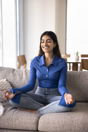 Serene Indian woman do meditation practice seated in lotus position on couch in living room, deeply...