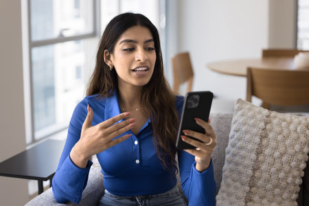 Young Indian woman sit on couch, holding smartphone engaged in video call or online conversation, speaking to friend or family member, explaining or discussing news. Apps usage, remote communicationの写真素材