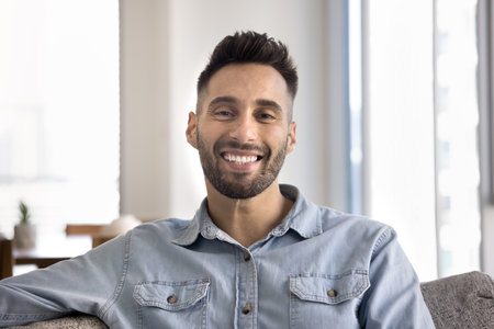 Young Hispanic man looking at camera with joyful expression seated on comfortable couch in modern...
