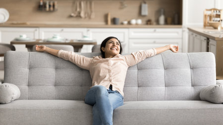 Pleasure to be homeowner. Happy carefree young girl recline on comfy couch relax stretch arms enjoy buying new furniture to modern house studio flat glad spending free weekend at clean own rented homeの写真素材