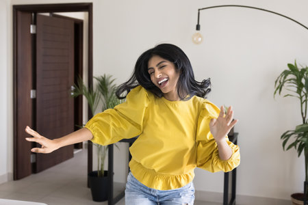 Young 30s energetic Indian woman dancing indoors, celebrate independence or relocation day to new...